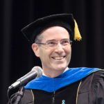 (Dean Christopher Long) Older man with glasses in academic robes with blue trim neck chevron