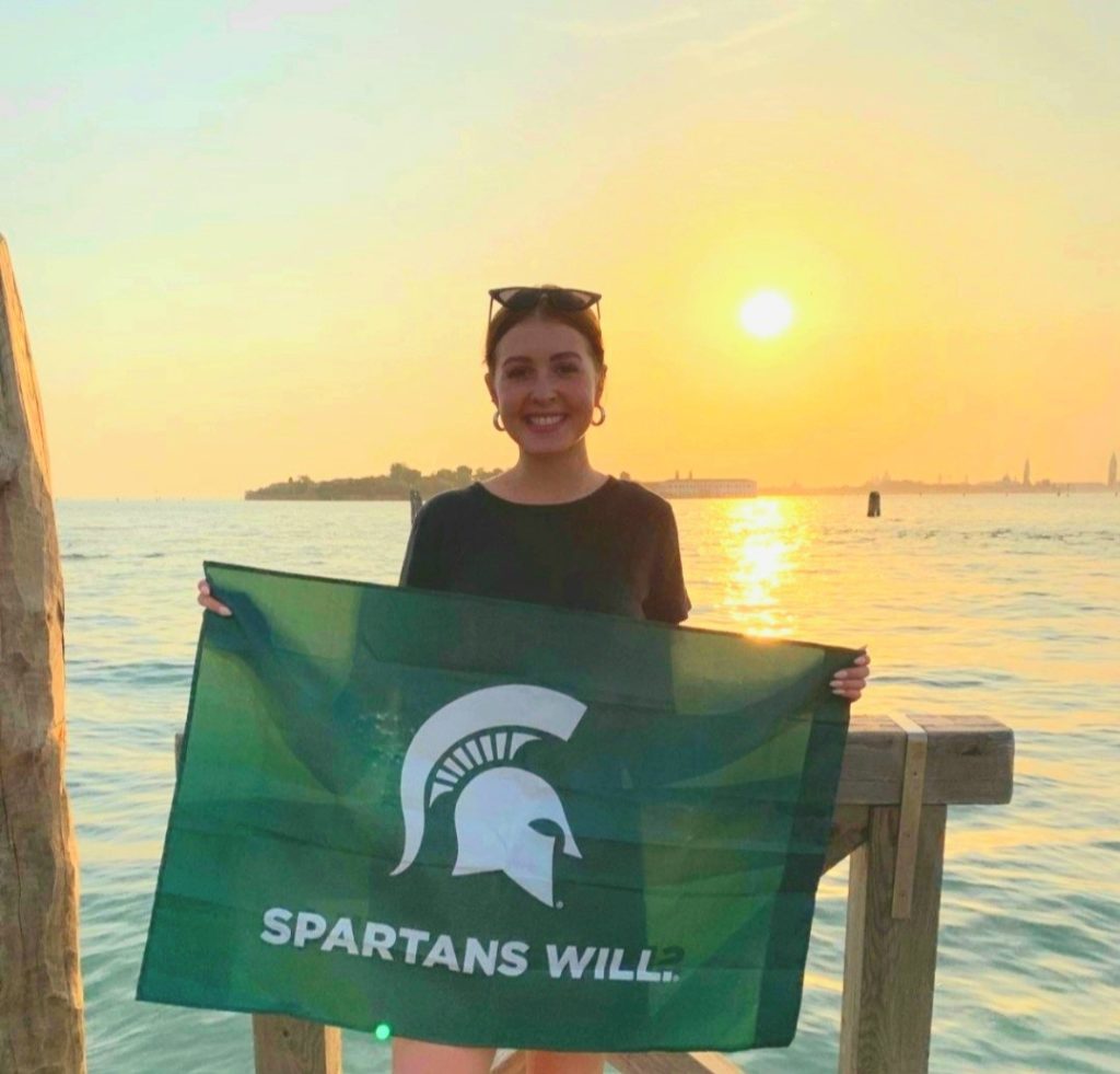 woman wearing a black top holding a Michigan State flag standing in front of a sunset above the water